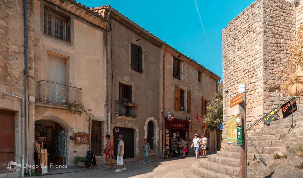 Calle soleada con torre de piedra, fachadas antiguas y peatones en el centro histórico.