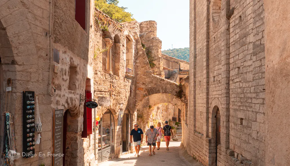 Calle estrecha de piedra con arcos, tiendas y visitantes caminando en Saint-Guilhem-le-Désert.