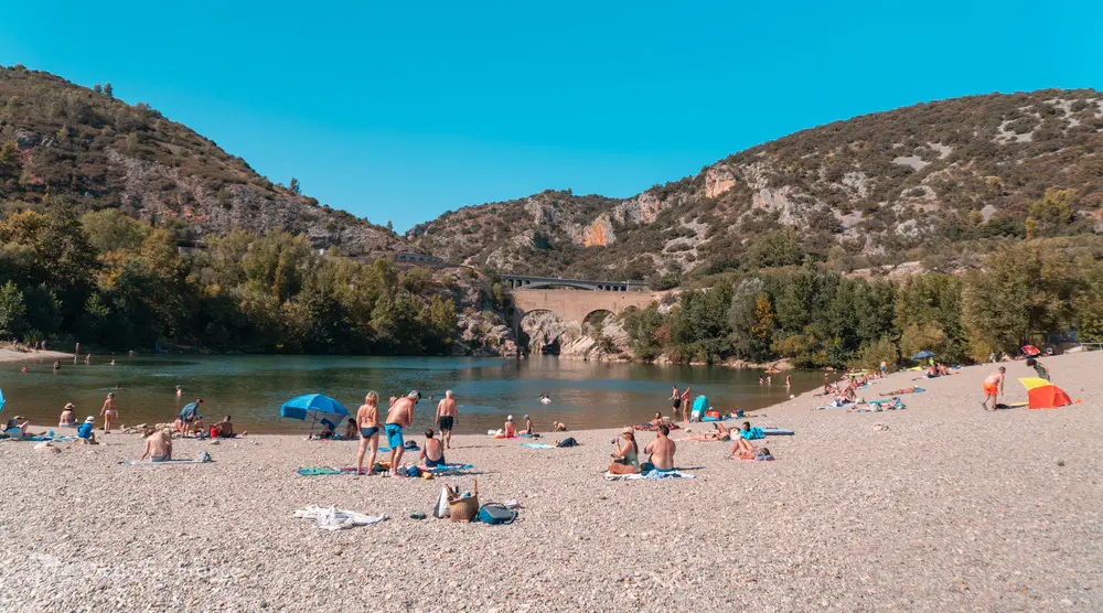 Playa de cantos rodados junto al río con bañistas y montañas alrededor