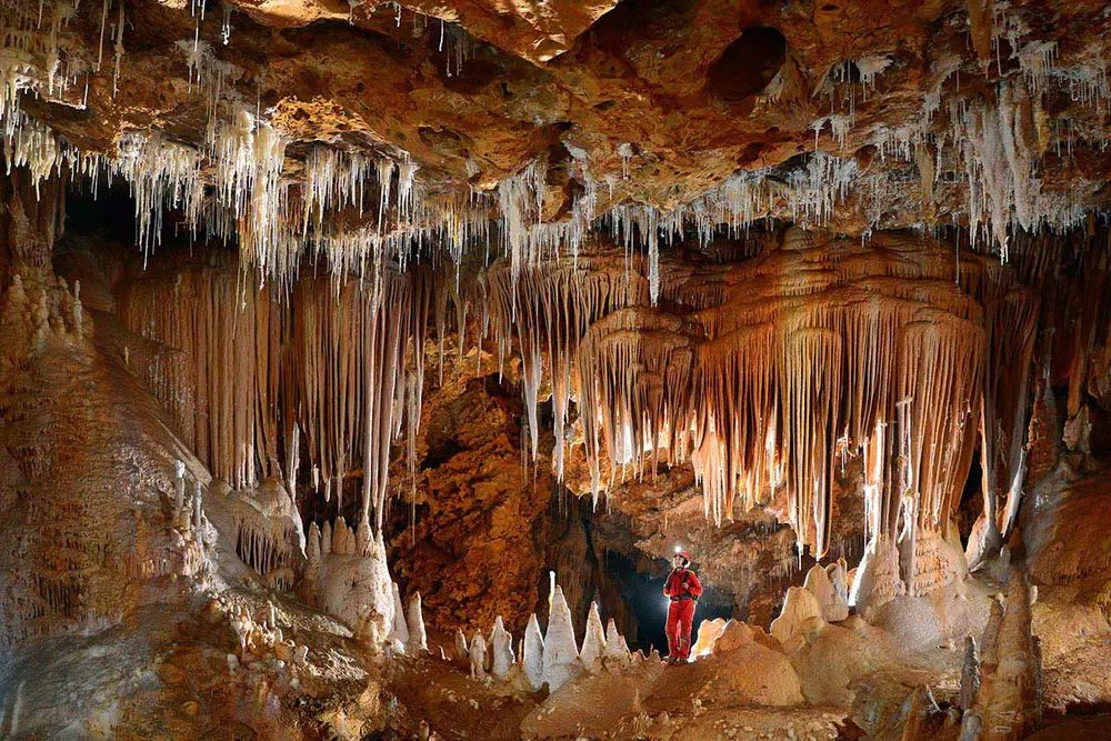 Sala subterránea con estalactitas, estalagmitas y visitante observando formaciones en una cueva