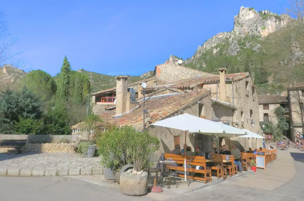 Terraza con mesas al aire libre frente a edificio de piedra en Saint-Guilhem-le-Désert, rodeado de colinas.
