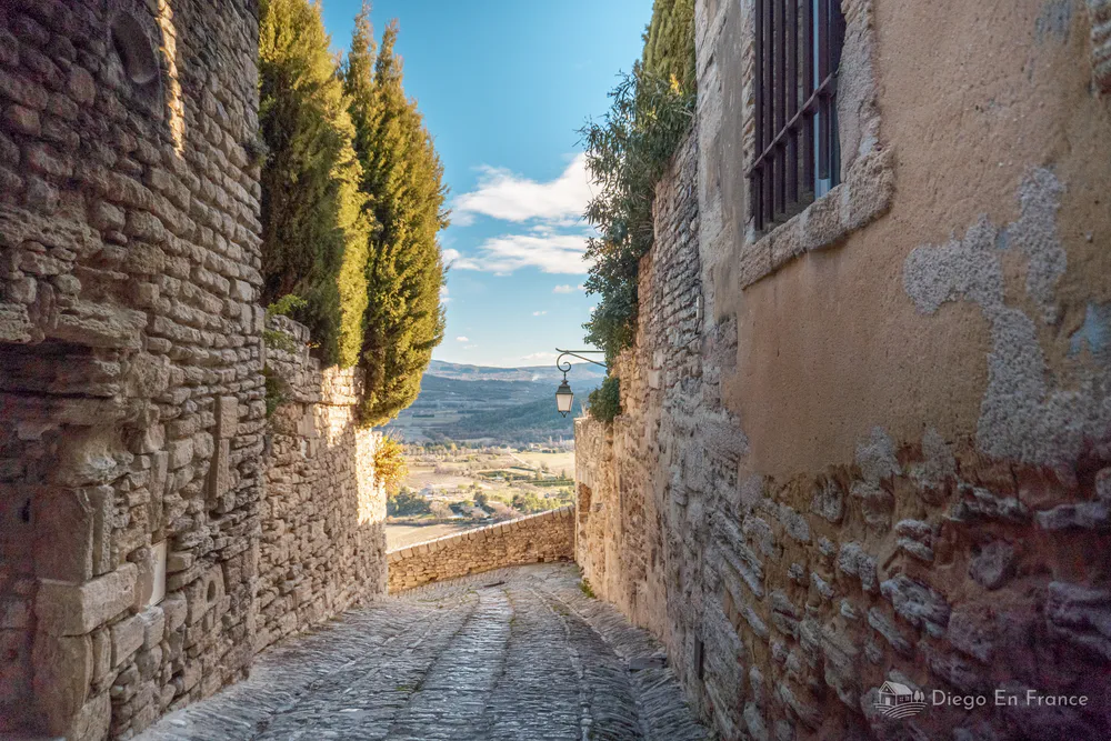 Charming cobblestone street in Gordes, Provence, with panoramic views of the Luberon valley, surrounded by stone walls and cypress trees.