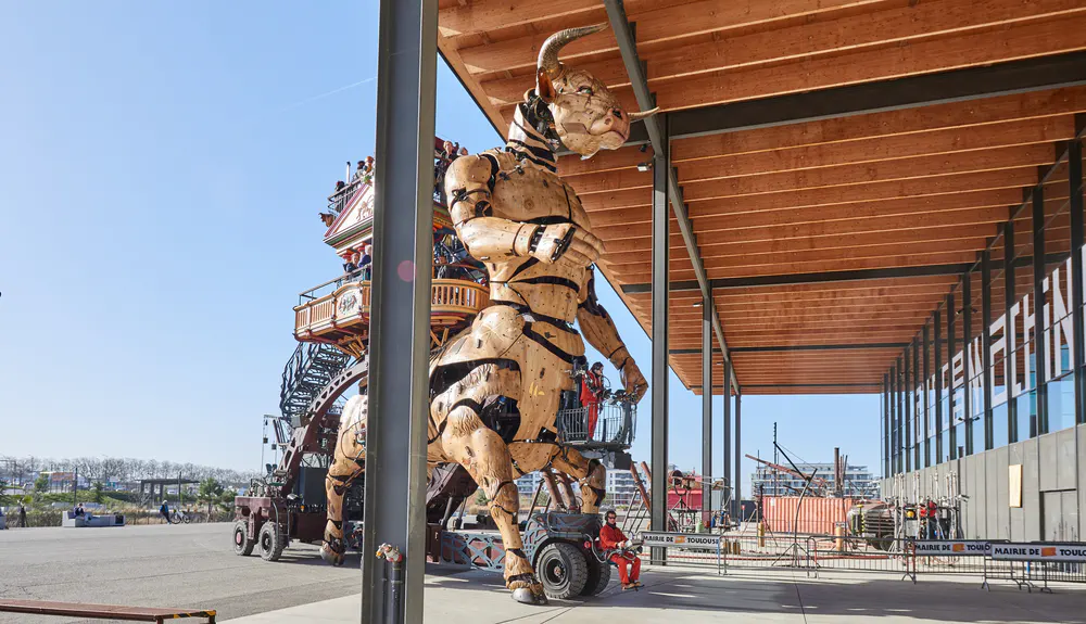 Giant wood and metal Minotaur in motion in front of La Halle de la Machine in Toulouse, with visitors on board and modern structure in the background. One of the best things to see in Toulouse