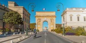Foto de una mujer en bicicleta con el arco del trinfo en Montpellier de fondo