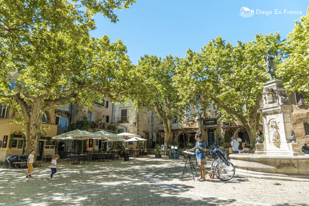 Square with a central statue, café terraces, and leafy trees in a village in southern France