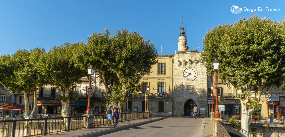 Bridge lined with trees and clock tower in the background in a village in the south of France
