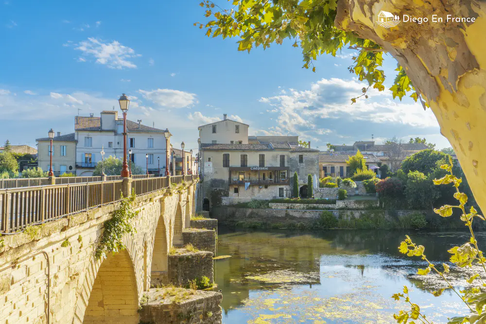 Stone bridge over a calm river with low houses and trees on both banks.