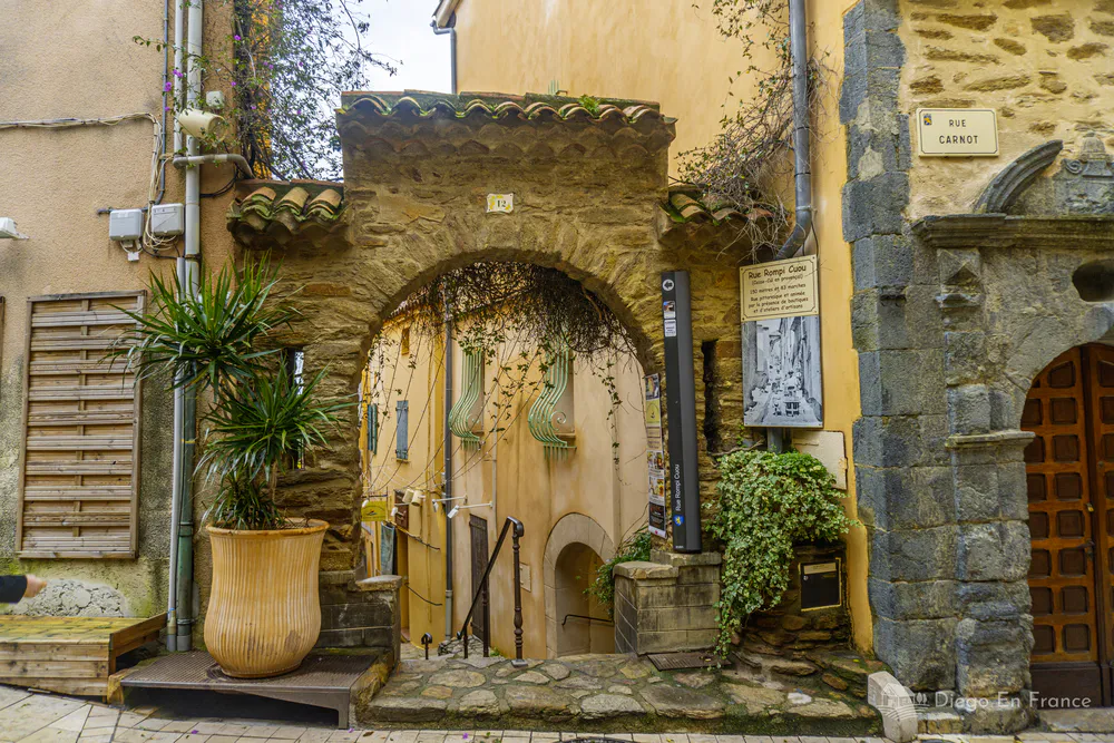 Arco de piedra con carteles informativos y vegetación en una calle estrecha