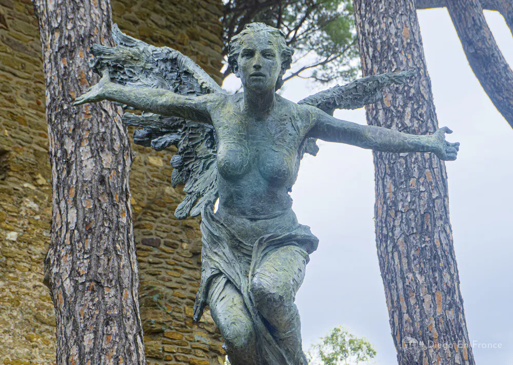 Escultura femenina de bronce situada entre pinos junto a una muralla de piedra