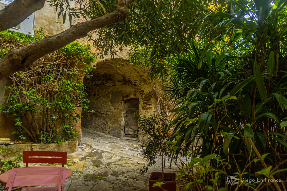 Pasaje de piedra con arco antiguo, vegetación densa y mesas al aire libre