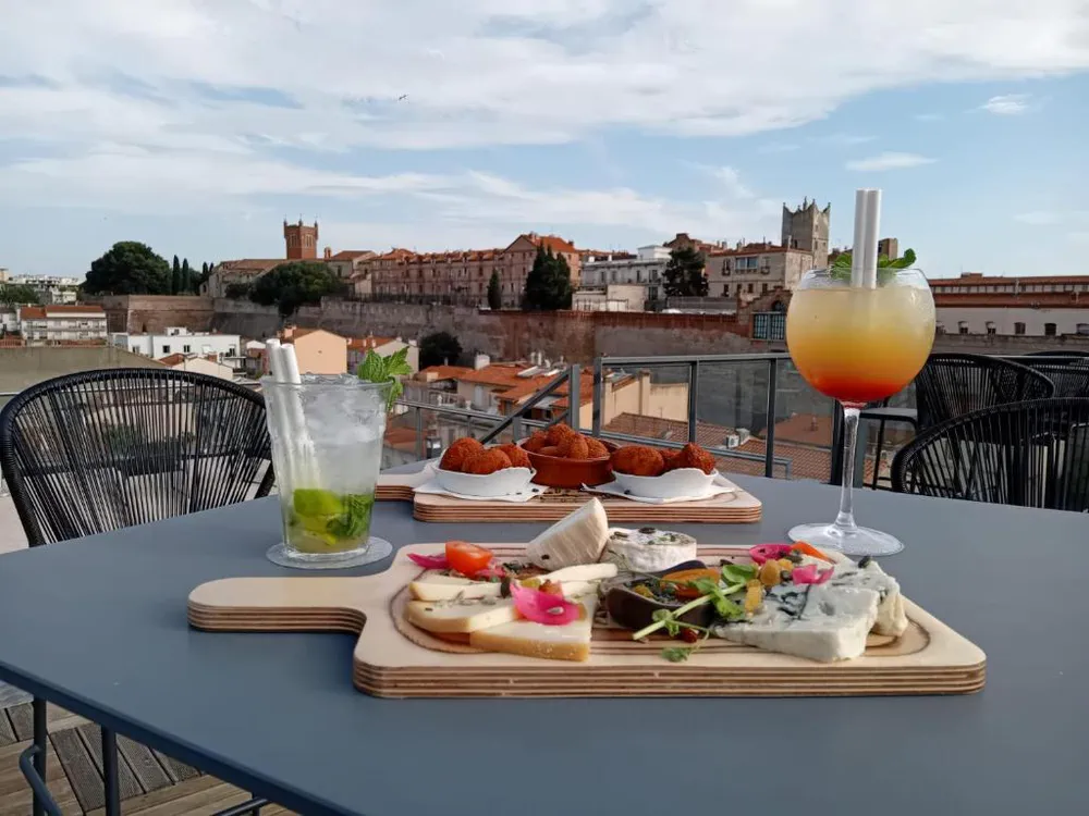 Mesa con aperitivos y tragos sobre una terraza con vista al centro histórico y monumentos de Perpiñán al atardecer.