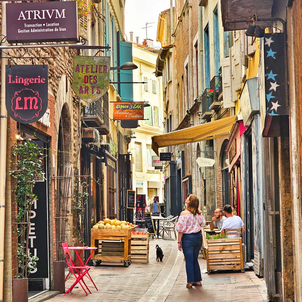 Calle peatonal estrecha en Perpiñán con tiendas locales, frutas frescas, arte y una mujer caminando entre terrazas acogedoras.
